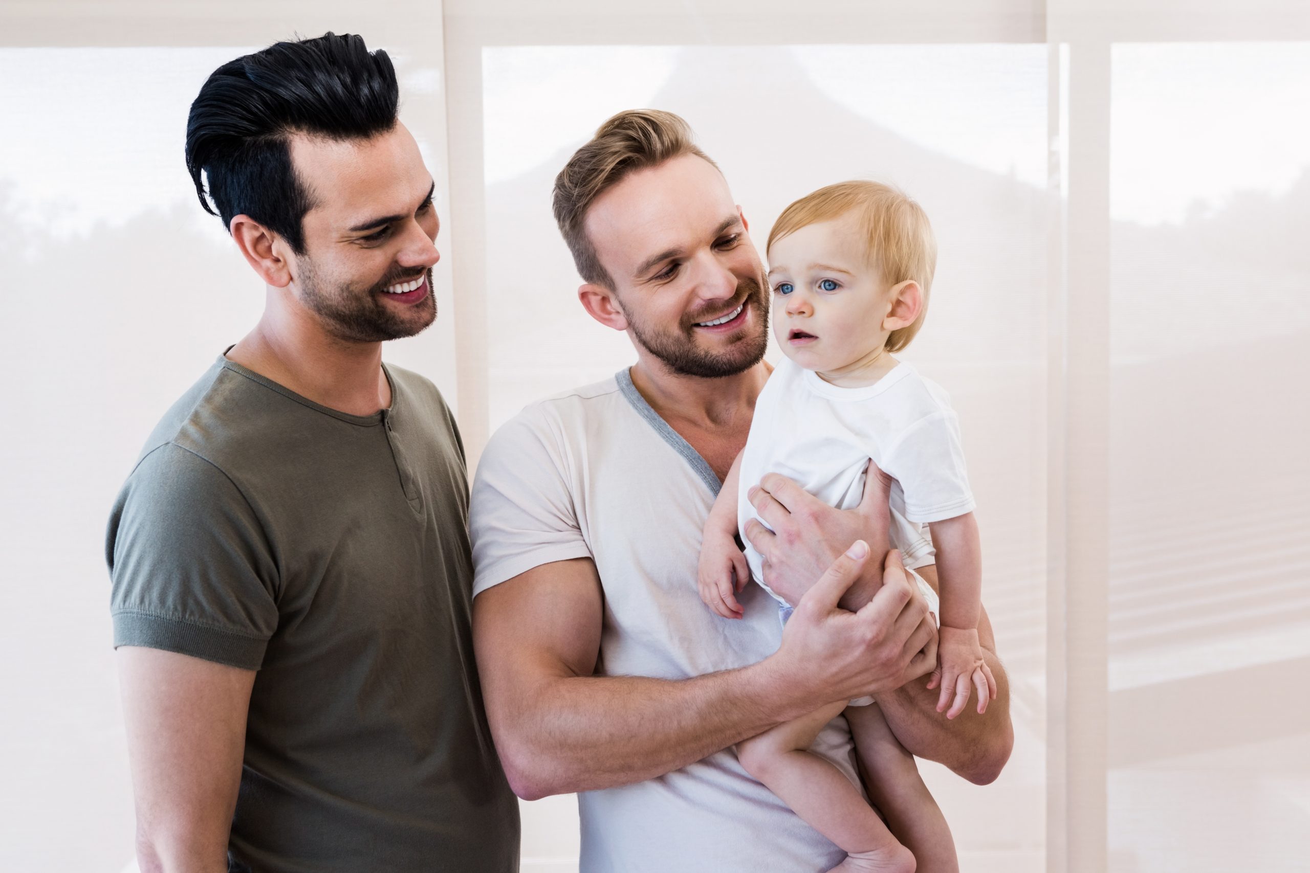 Couple standing side by side with one man holding baby. Both men smiling at baby