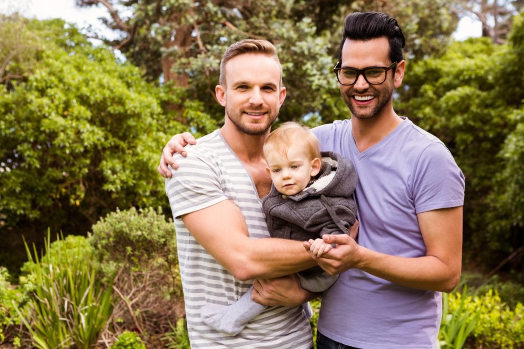 Couple holding their baby together and both men are smiling into the camera